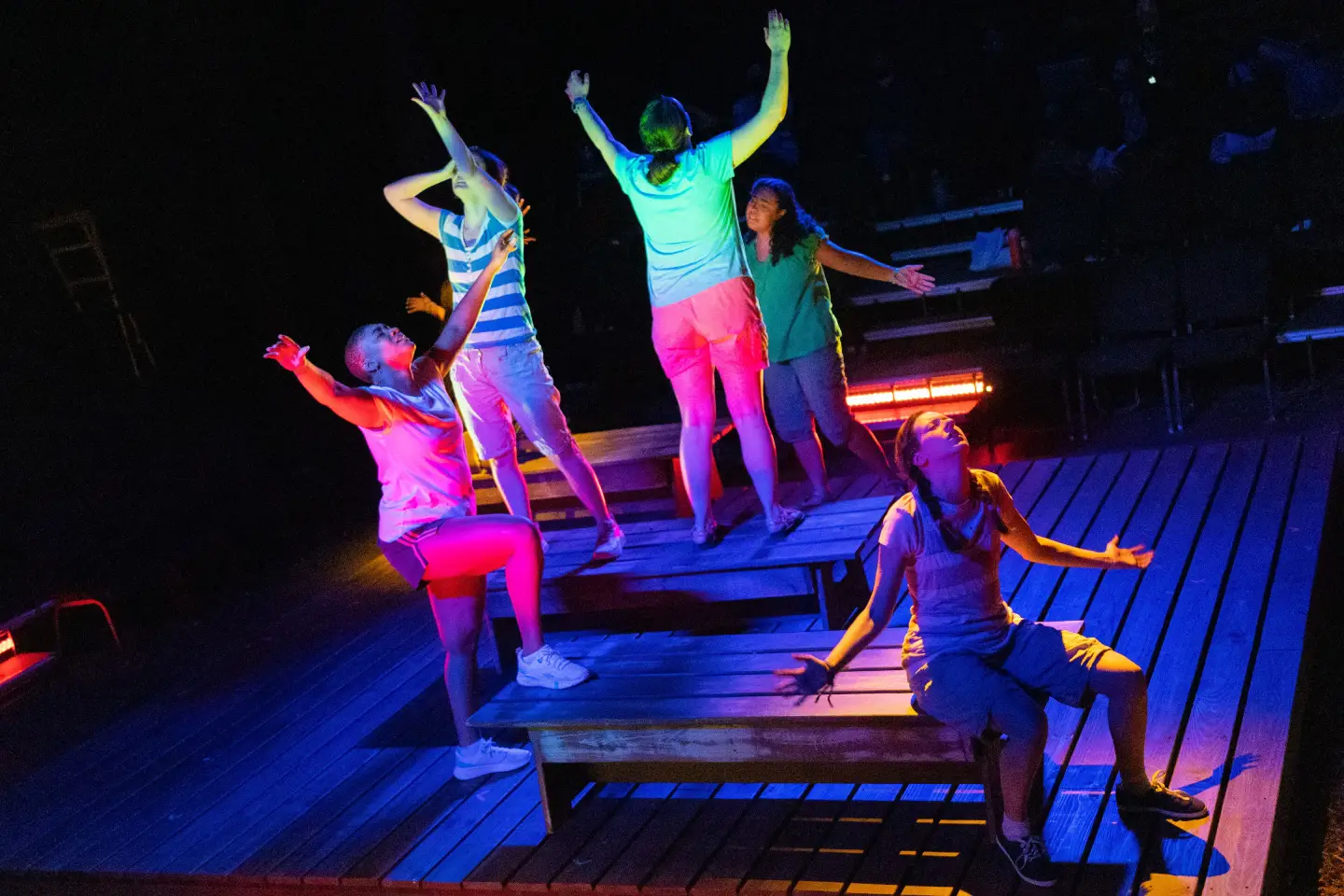 A group of young performers stand and sit on wooden risers under colorful stage lighting, arms raised.